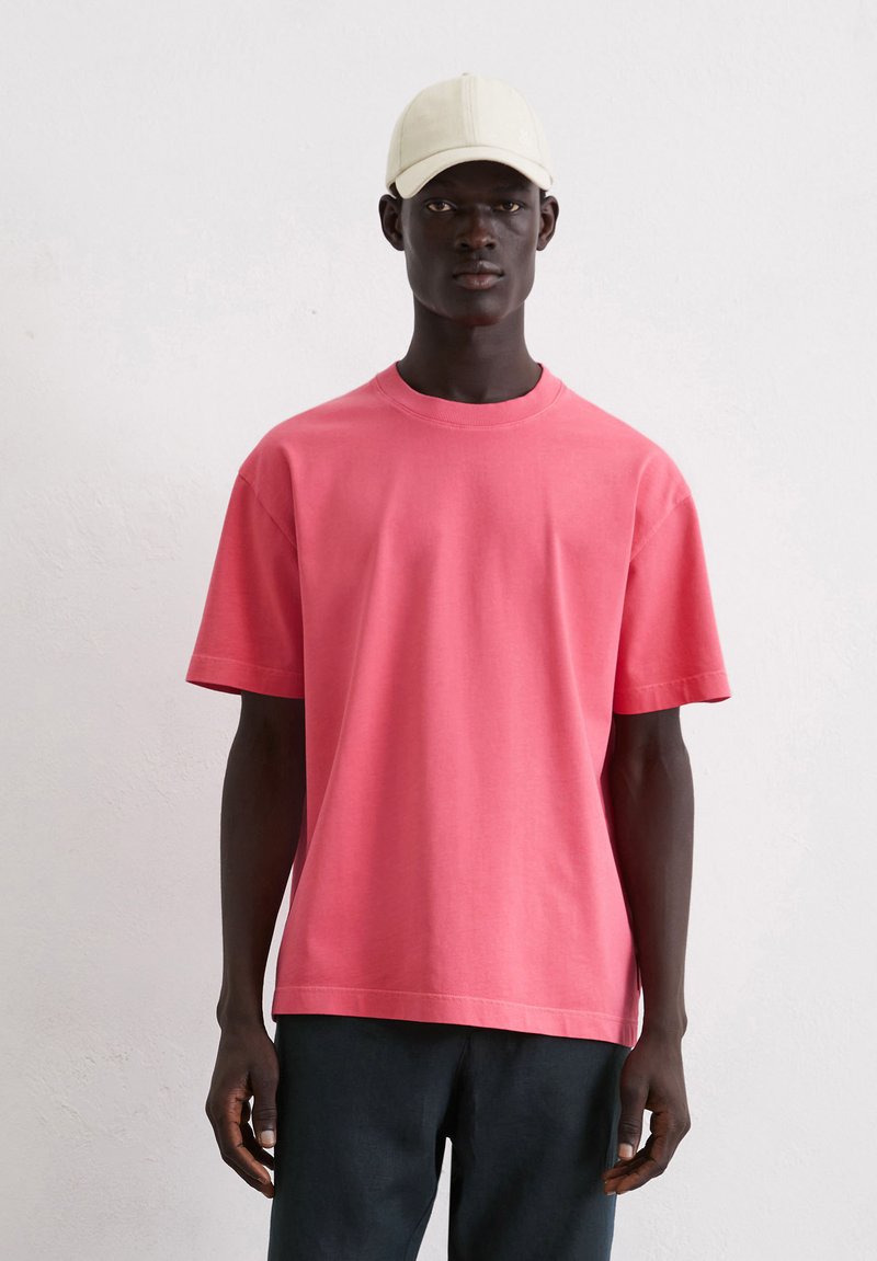 Korte mouwen, ronde hals t-shirt in fel koraal stof met een gladde textuur, gecombineerd met donkere broeken en een lichtgekleurde pet.
