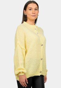 Cardigan en tricot jaune avec un col rond, de grands boutons et des poignets côtelés. Présente une coupe ample et un motif texturé.