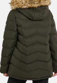 Veste d'hiver matelassée vert olive avec capuche bordée de fourrure, surpiqûres en chevron et écusson rond sur la partie supérieure de la manche gauche.