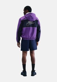Giacca Nike a blocchi di colore con design viola e blu navy, orlo elastico, cappuccio e grande logo sulla schiena. Abbinata a pantaloni corti scuri e calze.