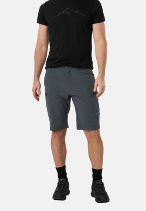 Mann trägt schwarzes T-Shirt, graue knielange Cargo-Shorts mit Reißverschlusstaschen, schwarze Socken und schwarze Sportschuhe, steht vor weißem Hintergrund.