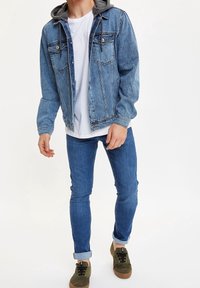 Chaqueta de mezclilla con capucha gris, color azul claro, dos bolsillos en el pecho y cierre de botones, combinada con jeans ajustados azules y zapatos verdes.