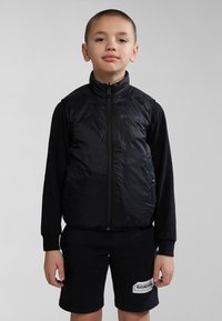 Doudoune sans manches légère noire avec fermeture éclair, col haut et détail du logo, portée sur une chemise noire à manches longues et un short.