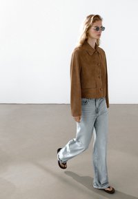 Giacca in suede marrone con colletto e bottoni, abbinata a jeans a gamba dritta azzurri chiari e sandali neri. La modella indossa occhiali da sole rotondi.