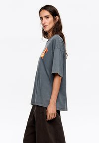 Szary, oversize t-shirt z krótkimi rękawami i okrągłym dekoltem, z pomarańczowym nadrukiem graficznym na przodzie. Miękki, teksturowany materiał.