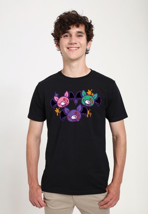 Giovane uomo con capelli ricci sorride indossando una t-shirt nera con facce di pipistrelli colorate in stile cartone animato con schizzi di vernice e ali su sfondo bianco.