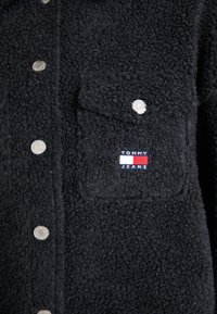 Jachetă din fleece neagră cu capse argintii, un buzunar pe piept și o etichetă cu logo în culorile roșu, alb și albastru. Material texturat.
