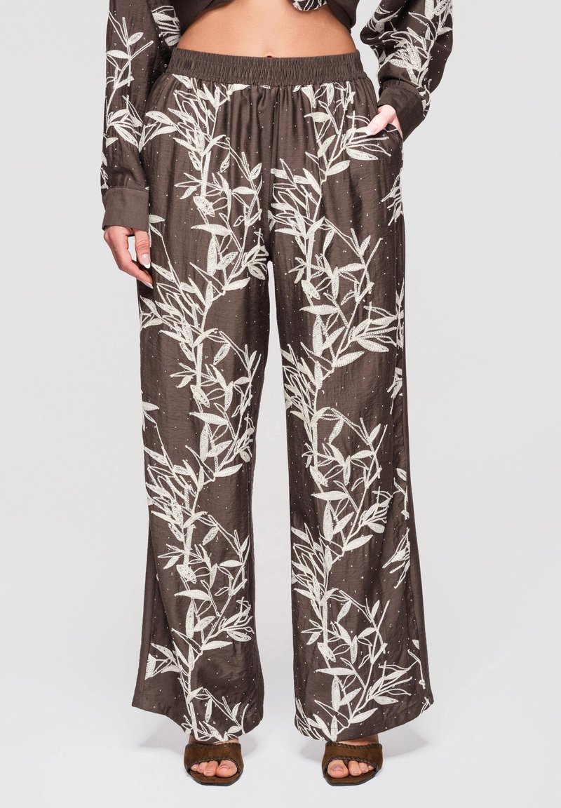 Pantalon marron à jambes larges avec motifs de vignes blanches feuillues, taille élastique et poches, porté avec des sandales marron à bout ouvert.
