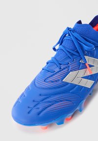 Blue soccer cleat with orange dot pattern, silver logo, blue laces, and orange studs on white background.