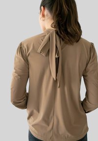 Vrouw met donker haar in een paardenstaart, gekleed in een beige blouse met lange mouwen en een strikje aan de achterkant van de hals, met gouden oorringen.