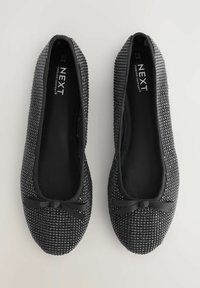 O pereche de balerini negri sclipitori cu fundițe mici pe vârfuri, etichetați "NEXT Forever Comfort", arătați din partea de sus pe un fundal alb.