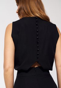 Czarny bezrękawnik z zaokrąglonym dekoltem z tyłu, z rzędem guzików pokrytych tkaniną oraz małym wycięciem z tyłu. Gładka tekstura.