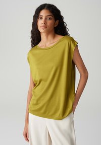 Top verde oliva a maniche corte realizzato in tessuto liscio, con una vestibilità rilassata e un ampio scollo rotondo, abbinato a pantaloni chiari.