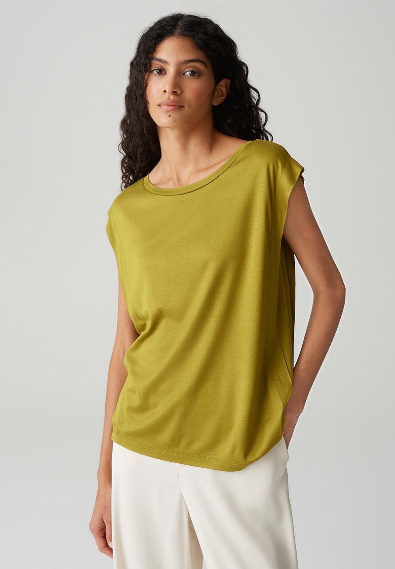 Top verde oliva a maniche corte realizzato in tessuto liscio, con una vestibilità rilassata e un ampio scollo rotondo, abbinato a pantaloni chiari.