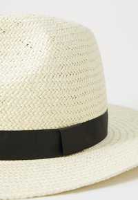 Sombrero de paja trenzada en color natural con una ancha banda de cinta negra. Presenta una copa redondeada y un ala ligeramente curvada.
