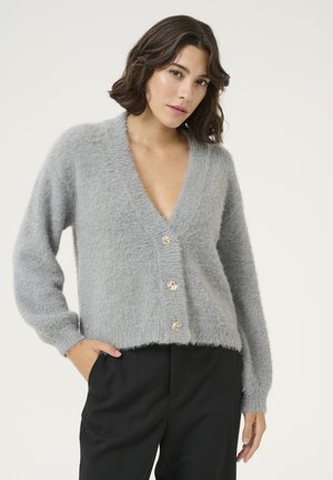 Grå cardigan, fluffig textur, djup V-ringning, långa ärmar, fem dekorativa knappar, kort passform, matchad med svarta byxor.