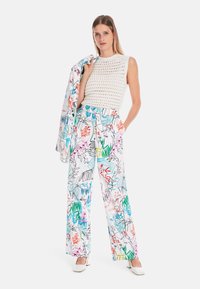 Häkel-gestricktes weißes Tanktop kombiniert mit weiten Hosen, die ein farbenfrohes Blumenmuster aufweisen. Die Modell hält eine passende leichte Jacke.