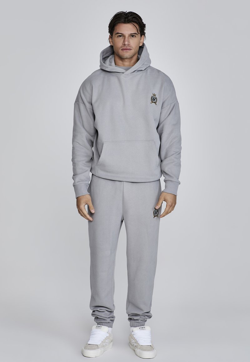Sudadera gris y joggers a juego de tejido suave. La sudadera cuenta con un bolsillo frontal y un emblema en el pecho. Las zapatillas tienen cordones blancos y detalles texturizados.