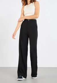 Femme portant un haut sans manches blanc, un pantalon noir taille haute à jambes larges et des baskets noir et blanc, se tenant devant un fond uni.