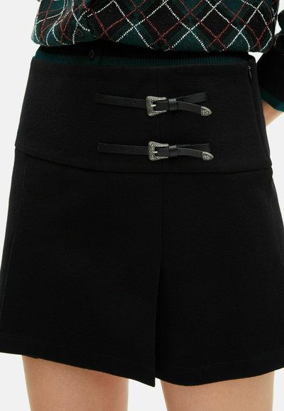 Čierna sukňa s vysokým pásom, ktorá má dve dekoračné kožené remeňové pásky so striebornými sponami, nosená s tmavozeleným a červeným vrchom s károvaným vzorom.