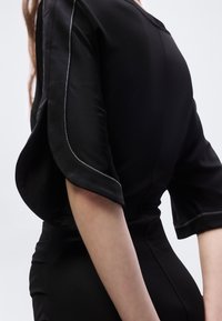 Κοντινό πλάνο ενός ατόμου που φοράει μαύρο φόρεμα με φαρδιά, κυματιστά μανίκια που έχουν άσπρη ραφή κατά μήκος των άκρων.