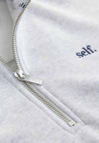 Šedá mikina na zip s kovovým zipem stříbrné barvy a brandovaným poutkem na zip, na látce modře vyšitý nápis "self.". Měkká textura.