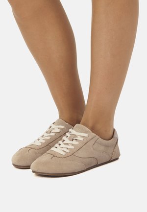 Piedi che indossano scarpe casual beige in suede con lacci bianchi su uno sfondo bianco, mostrando le gambe inferiori e le caviglie.