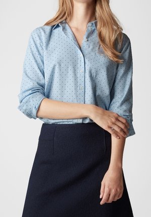 Vrouw draagt lichtblauw, lang mouw, knoophemd met kleine stippen en een donkerblauwe rok, armen gekruist ter hoogte van de taille, tegen een effen achtergrond.