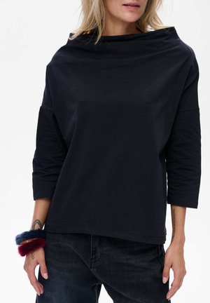 Vrouw in een losse zwarte lange mouwtop met een brede halslijn, zwarte broek en fuzzy rode en blauwe armbanden om haar linkerpols.