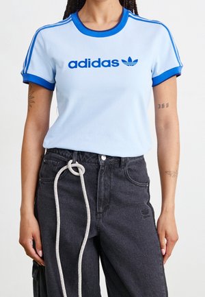 Person trägt ein hellblaues Adidas-T-Shirt mit blauen Besätzen und Streifen, kombiniert mit hoch taillierten schwarzen Jeans und einem weißen Seilgürtel, der zu einem Knoten gebunden ist.