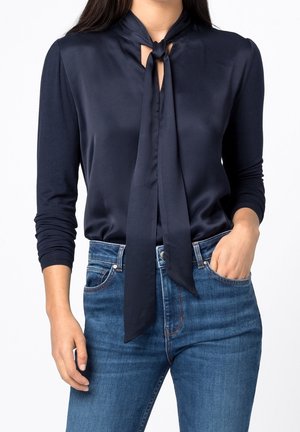 Mujer vestida con una blusa de manga larga color azul marino con detalle de nudo delantero y jeans de mezclilla azul, con una mano en el bolsillo, sobre un fondo blanco.