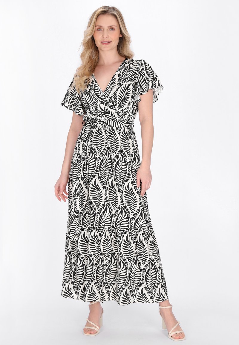 Blonde Frau, die ein schwarz-weißes, blattgemustertes Wickelkleid mit Flatterärmeln und weißen Sandalen mit Absatz trägt, steht vor weißem Hintergrund.