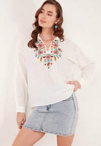 Blusa bianca a maniche lunghe con ricami floreali colorati sul colletto, abbinata a una gonna di jeans azzurri. Presenta una vestibilità morbida.