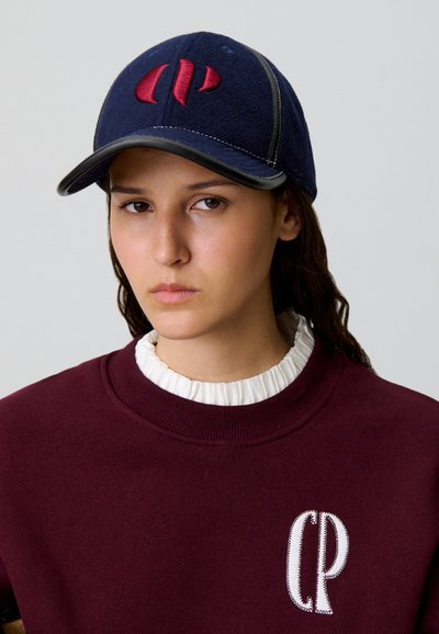 Giovane che indossa un cappello blu navy con ricami rossi e una felpa bordeaux con un logo bianco "CP" sopra un colletto bianco arricciato.
