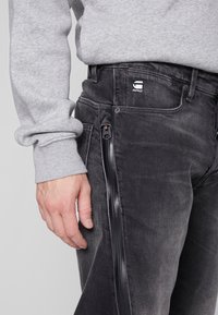 Jeans in denim nero con una finitura sbiadita, zip laterale e accento con logo sulla cintura, abbinati a una felpa a maniche lunghe con polsini di colore grigio chiaro.