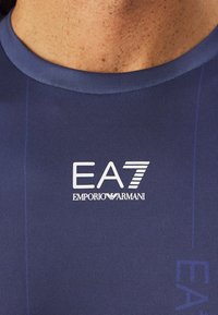 Camiseta deportiva azul marino con superficie texturizada, con un logo blanco "EA7" y "EMPORIO ARMANI" en el pecho, escote redondo.