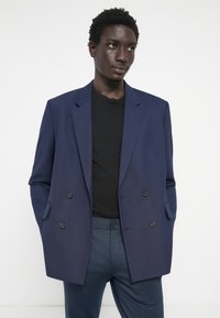 Blazer croisé bleu marine avec deux boutons noirs et revers crantés, porté sur un t-shirt noir, associé à un pantalon bleu foncé.