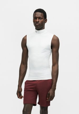 Hombre de pie con camiseta blanca sin mangas de cuello alto y pantalones cortos granate sobre un fondo claro liso.