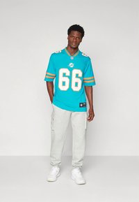 Turkio spalvos NFL marškinėliai su oranžiniais akcentais ir numeriu 66, derinami su pilkomis kargo sportinėmis kelnėmis ir baltais sportbačiais.