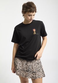 Joven mujer vestida con una camiseta gráfica negra y una falda de estampado de leopardo, mirando hacia abajo, con cabello corto castaño y pendientes de aro, frente a un fondo blanco.