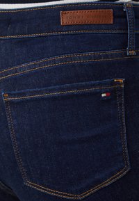 Jeans de mezclilla azul oscuro con costuras naranjas, que presentan una etiqueta de cuero con el logo "TOMMY HILFIGER" y un pequeño acento de bandera en el bolsillo trasero.