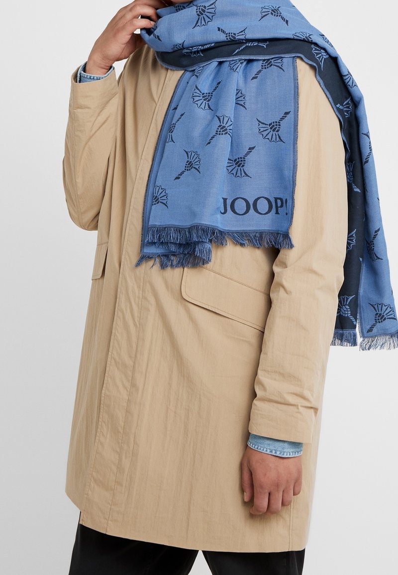 Trench-coat beige avec grande poche, porté sur une chemise en jean, accompagné d'une écharpe bleue à motifs portant le texte "JOOP!" et des bords frangés.