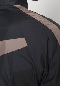 Svart jacka med en slät tekstur, med ljusbruna diagonala paneler längs axlarna och ryggen för kontrast.