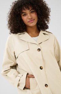 Beige knopenjas met een klassieke kraag, met een gestroomlijnd ontwerp en een voorzak, gemaakt van een soepele stof met een subtiele textuur.