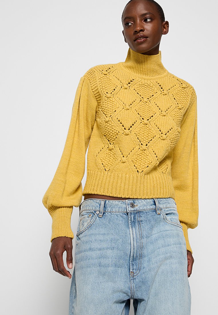 Donna che indossa un maglione giallo a collo alto con maniche a sbuffo e jeans chiari a vita alta, mentre guarda di lato.