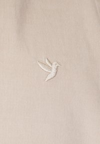 Beige stof med et tekstureret ribdesign, med et lille hvidt broderet kolibri-logo placeret på venstre side.