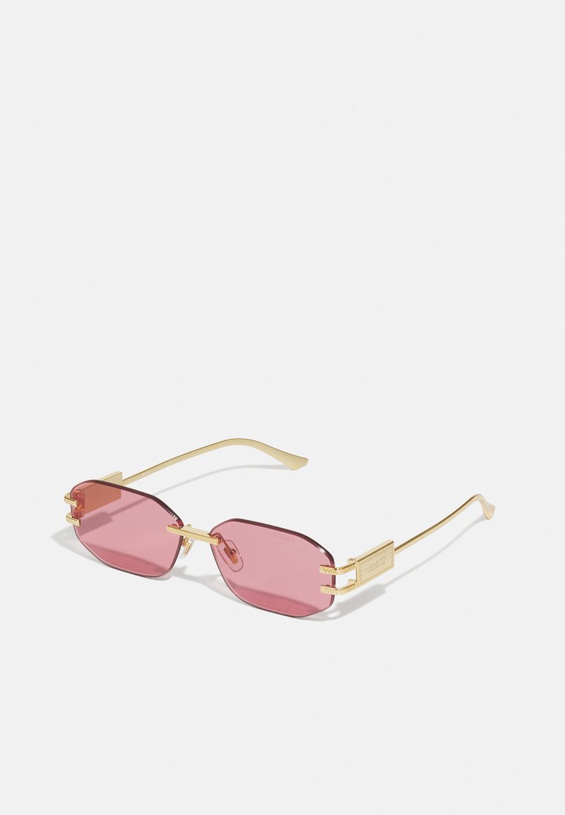Ochelari de soare octogonali cu nuanță roz, cu ramă din metal de culoare aurie și accente decorative pe laterale. Design ușor, cu brațe subțiri.