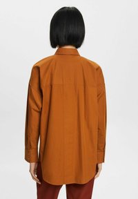 Camisa marrón de manga larga con un ajuste relajado, aberturas laterales y puños abotonados. Tejido de algodón suave con un detalle de costura trasera recta.