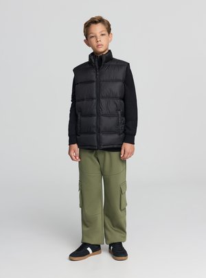 Czarna kamizelka puchowa z wysokim kołnierzem i poziomym pikowaniem. W zestawie z oliwkowymi spodniami cargo oraz czarnymi tenisówkami z białymi paskami.