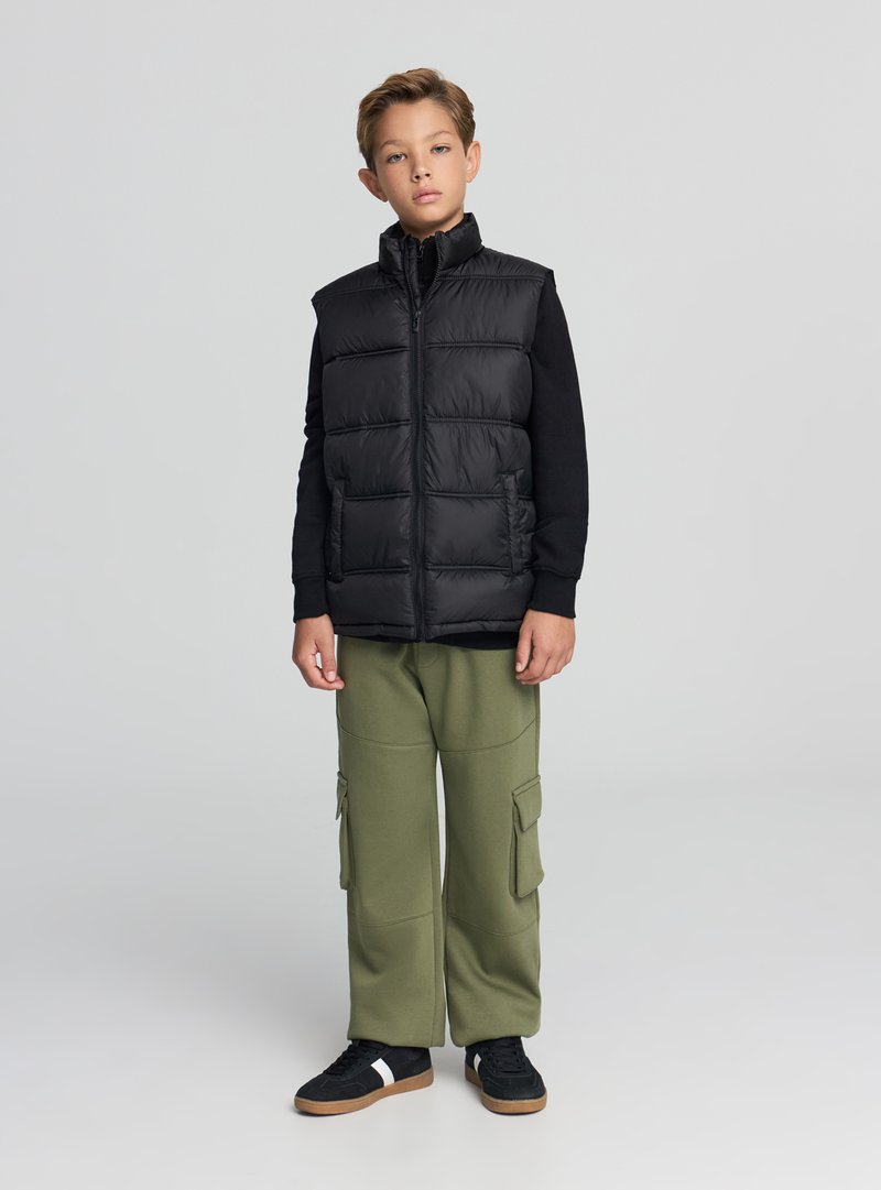 Czarna kamizelka puchowa z wysokim kołnierzem i poziomym pikowaniem. W zestawie z oliwkowymi spodniami cargo oraz czarnymi tenisówkami z białymi paskami.